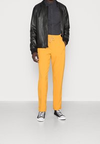 Muž oblečený v černé kožené bundě, tmavě šedé košili na knoflíky, jasně žlutých kalhotách s pružným pasem a černých vysokých teniskách stojící na bílém pozadí.