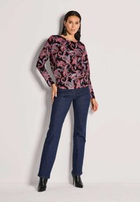 Langärmlige Bluse mit schwarzem Hintergrund und Paisleymuster in Rottönen und Blau, kombiniert mit dunkelblauen Jeans und schwarzen Stöckelschuhen.