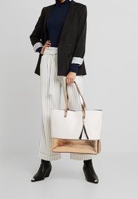 Bolsa tote con una parte superior blanca y un fondo metálico color rosa dorado. Incluye dos asas y un detalle en negro en el centro.