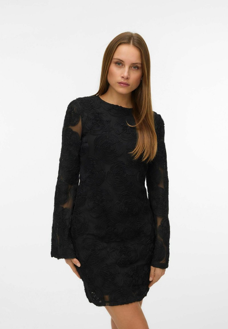 Femme portant une robe noire ajustée avec des motifs floraux texturés et des manches longues transparentes, debout devant un fond blanc uni.