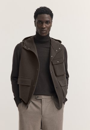 Mann trägt eine dunkelbraune ärmellose Jacke über einem schwarzen Rollkragenpullover und hellbraune Hose, steht mit den Händen in den Taschen.