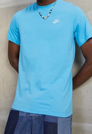 Person trägt hellblaues Nike-T-Shirt und Patchwork-blaue Shorts, mit einer Perlenkette, die einen weißen Gänseblümchenanhänger zeigt.