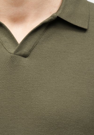 Gros plan d'une personne portant un polo texturé vert olive avec un col ouvert et sans boutons visibles.