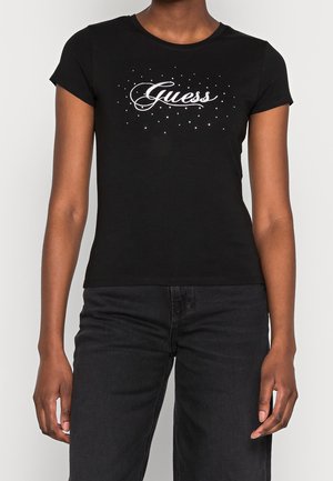 Femme portant un t-shirt noir ajusté avec le logo blanc "Guess" et de petits points décoratifs, assorti à un jean noir sur un fond uni.