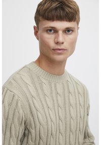 Pull en tricot beige avec un col rond et un motif texturé. Le tissu a l'air doux, avec des motifs torsadés en relief sur les manches.
