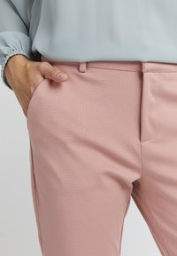 Růžové strukturované kalhoty s hladkým povrchem, s bočními kapsami a přiléhavým pasem. V kombinaci s světle modrou blůzou se elastickými manžetami.