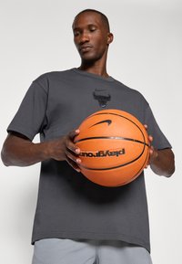 Grå Nike t-skjorte med en svart Bulls-logo, holdt av en person med en oransje basketball, glatt tekstur, avslappet passform.