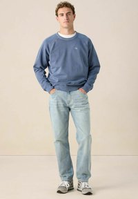 Jeune homme se tenant avec les mains dans les poches, portant un sweatshirt bleu délavé, un jean bleu clair et des baskets grises, sur un fond uni.