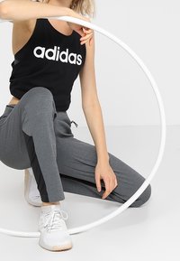 Czarny bezrękawnik z białym logo "adidas"; szare spodnie sportowe z czarnymi akcentami po bokach; białe buty sportowe; okrągły rekwizyt w pobliżu.