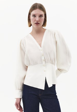 Femme portant un chemisier blanc à col en V, manches longues bouffantes et détails de boutons, associé à un jean bleu foncé, sur un fond blanc uni.