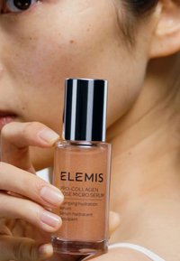 Glazen fles met een zilveren dop, gevuld met lichtbruin serum. Etiket leest "ELEMIS PRO-COLLAGEN ROSE MICRO SERUM" met tekst in het Engels en het Frans.