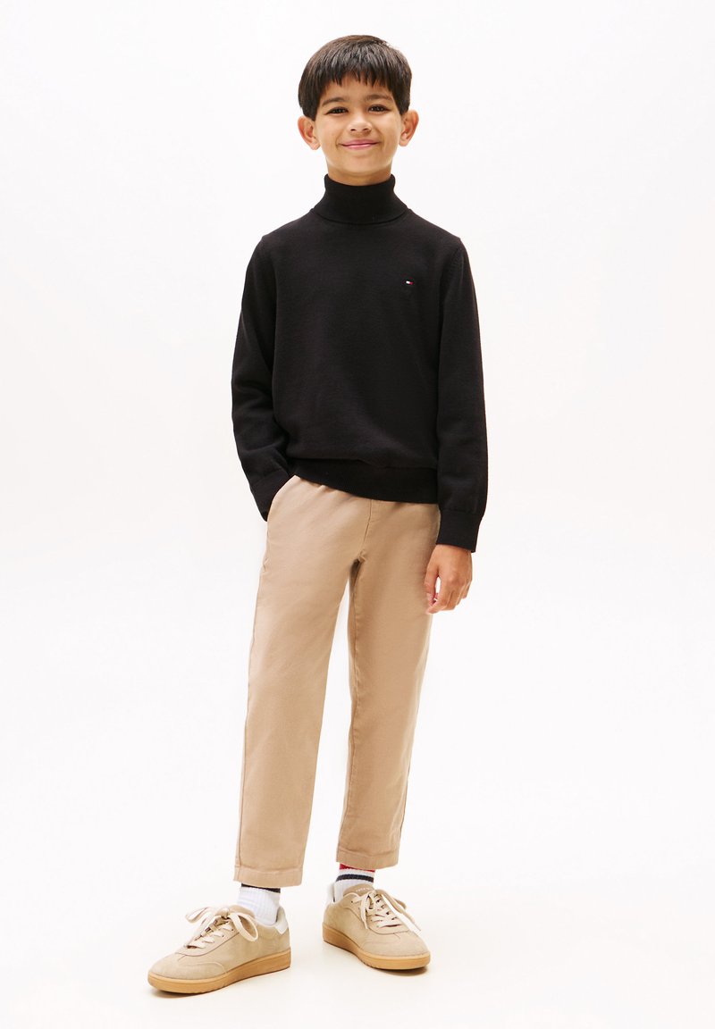 Maglione nero a collo alto, chinos beige e sneakers beige. Il maglione ha un piccolo logo sul petto; i pantaloni hanno un design a gamba dritta.