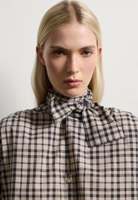 Beige and black checkered blouse with a high collar and bow tie. Features a button closure and smooth fabric texture.