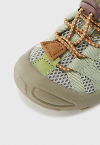 Κοντινό πλάνο από πεζοπορικό παπούτσι σε ανοιχτό πράσινο και καφέ, με υφασμάτινη δίχτυ, σχέδιο κορδονιών σε πορτοκαλί χρώμα και υφή στην ελαστική σόλα.