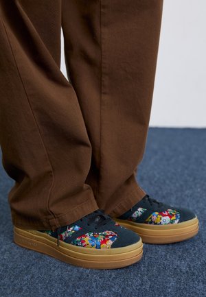 Pieds portant des baskets noires avec des motifs floraux colorés et des semelles épaisses couleur beige, associés à un pantalon marron ample sur un tapis bleu.