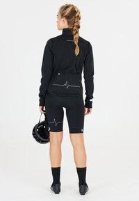 Zwarte fietsjack met een hoge kraag en een hartslaggrafiek op de achterkant, gecombineerd met zwarte shorts en fietsschoenen, terwijl hij een helm vasthoudt.