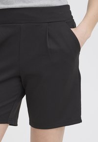 Detailný záber osoby oblečenej v čiernych krátkych nohaviciach s rukou nenútene vloženou vpravo do vrecka, viditeľný pás a predné záhyby.