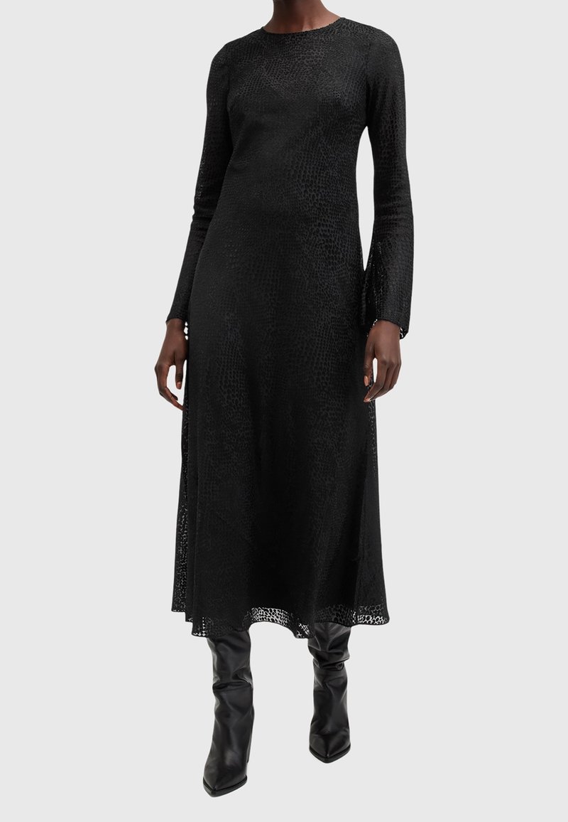 Femme portant une robe midi texturée noire à manches longues associée à des bottes noires montantes à bout pointu, debout sur un fond clair.