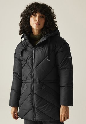 Kobieta o kręconych włosach, ubrana w czarną pikowaną zimową kurtkę z kapturem, nałożoną na oliwkowozielony golf.