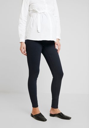 Persona embarazada que lleva una blusa blanca con lazo al frente, pantalones de maternidad ajustados negros y zapatos planos negros sin cordones, de pie sobre un suelo gris.