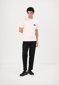 Camiseta de algodón rosa claro con mangas cortas y un logotipo de diamante negro en el lado izquierdo del pecho, combinada con pantalones negros y zapatillas negras.