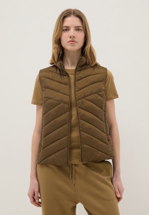 Jeune femme portant une veste matelassée sans manches marron par-dessus une chemise à manches courtes assortie et un pantalon à cordon, debout devant un fond uni.