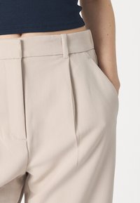 Beige op maat gemaakte broek met een gladde textuur, voorzien van voorplooien en zijzakken. De tailleband heeft riemlussen en een nette afwerking.