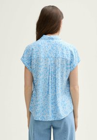 Camicia azzurra a maniche corte con motivo floreale, caratterizzata da una piega sul retro e un orlo rotondo. Tessuto liscio con una vestibilità rilassata.