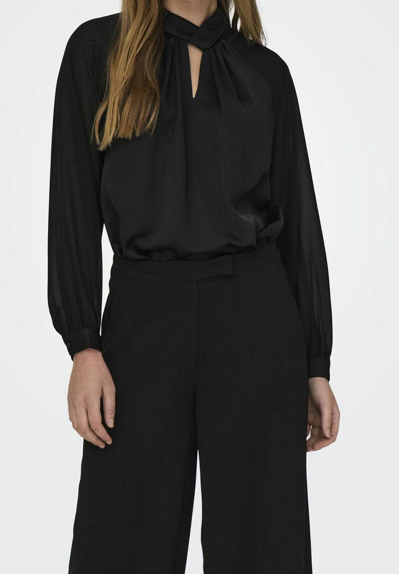 Femme portant un chemisier noir à manches longues avec détail d'encolure en trou de serrure et un pantalon noir taille haute à jambes larges sur un fond uni.