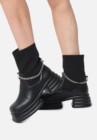 Svarta läderankelboots med ribbat svart strumpdetalj, kraftig klack och silverkedjeaccent. Texturerad tyg kontrasterar med slät läder.
