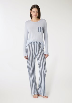 Młoda kobieta stoi boso, ubrana w jasnoniebieską bluzkę z długim rękawem z paskowaną kieszenią na piersi oraz pasujące szerokie spodnie w paski.