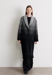 Cappotto lungo con gradiente dal nero al grigio, realizzato in materiale peloso. Presenta un ampio colletto e due tasche frontali. L'orlo ha un dettaglio frangiato.