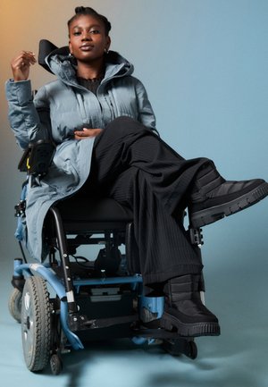 Gepolsterter grauer Mantel mit hohem Kragen, schwarze gerippte Hose und klobige schwarze Stiefel. Der Rollstuhl hat einen blauen Rahmen und doppelte Räder.