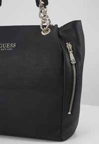 Zwarte leren Guess handtas met zilveren kettingband, zijrits vak en verdiept merknaam met "EST 1981" aan de voorkant.