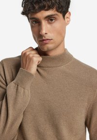 Młody mężczyzna z krótkimi kręconymi włosami, ubrany w beżowy sweter z wysokim kołnierzem, trzymający kołnierz jedną ręką, patrzy przed siebie.