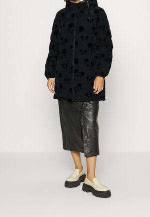 Frau trägt eine schwarze Steppjacke mit Mickey-Mouse-Aufdruck, einen schwarzen Leder-Midirock und beige Plateau-Loafer und steht vor einem weißen Hintergrund.