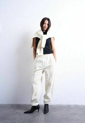 Ung kvinde iført hvide bukser og en sweater over en sort ærmeløs top, stående med hænderne i lommerne mod en hvid væg.