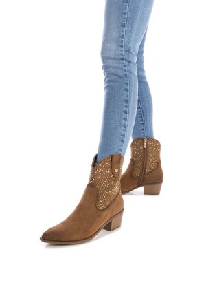 Botas de tobillo de ante marrón con adornos dorados, combinadas con unos pantalones vaqueros ajustados de un azul claro, mostradas sobre un fondo blanco.