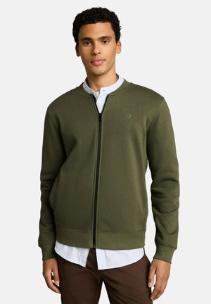 Junger Mann trägt eine olivgrüne Reißverschlussjacke über einem weißen Hemd und braunen Hosen, stehend vor einem schlichten hellen Hintergrund.