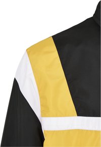Épaule de veste avec un design color-block en sections de tissu noir, jaune et blanc et détails de couture visibles.