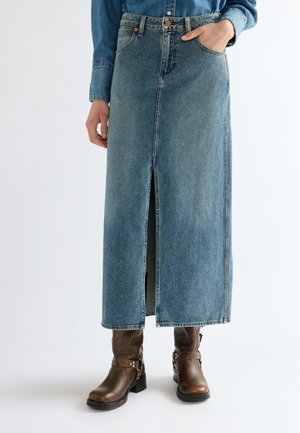 Personne portant une longue jupe en jean bleu avec une fente à l'avant et des bottines en cuir marron, main dans la poche de la jupe.