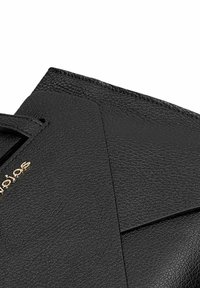 Close-up van een zwart lederen handtas met textuur, waarbij de voorflap en kleine gouden merkletters in de linkerbovenhoek zichtbaar zijn.