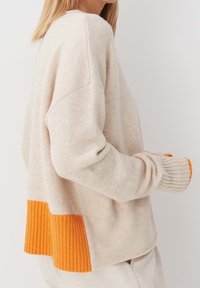 Beżowy sweter z dzianiny z prążkowanym pomarańczowym dolnym fragmentem i pomarańczowymi akcentami na rękawach. Posiada luźny krój i projekt z opadającymi ramionami.