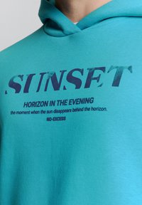 Turquoise hoodie met blauwe tekst "SUNSET," met een zachte textuur en een kangaroo zak. De tekst bevat citaten over de avond.