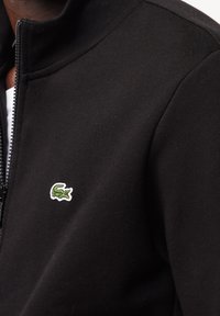 Giacca nera con zip e logo di coccodrillo ricamato verde sul lato sinistro del petto, indossata sopra una camicia bianca.