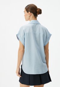 Camisa de manga corta azul claro con cuello, tela texturizada y puños enrollados, combinada con una falda plisada oscura de rayas.