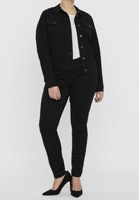 Persoon in een zwarte denimjack, zwarte broek, witte top en zwarte hoge hakken staande tegen een effen achtergrond.