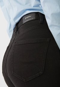 Jeans negros de mezclilla con una textura suave, que cuentan con un bolsillo trasero y una etiqueta de marca en la cinturilla, combinados con una camisa de color azul claro.