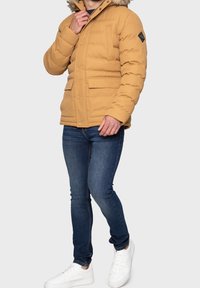 Doudoune beige avec capuche doublée de fourrure, dotée de deux poches avant et d'un écusson sur la manche. Portée avec un jean foncé et des baskets blanches.