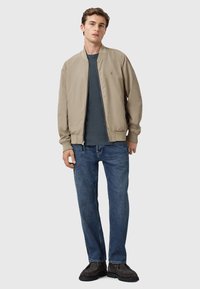 Chaqueta bomber de color beige claro con puños acanalados, llevada sobre una camiseta negra. Combinada con jeans azules y zapatos oscuros casuales. Diseño sencillo.